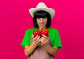 Free photo surprised young female gardener in uniform wearing gardening hat holds and looks at red peppers