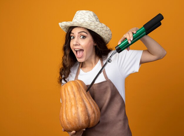 Surprised young female gardener in uniform wearing gardening hat holds pumpkin and garden clippers isolated on orange wall with copy space