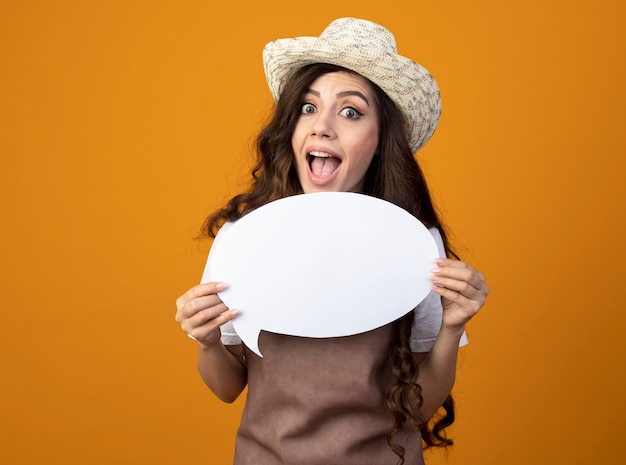 Surprised young female gardener in uniform wearing gardening hat holds speech bubble isolated on orange wall