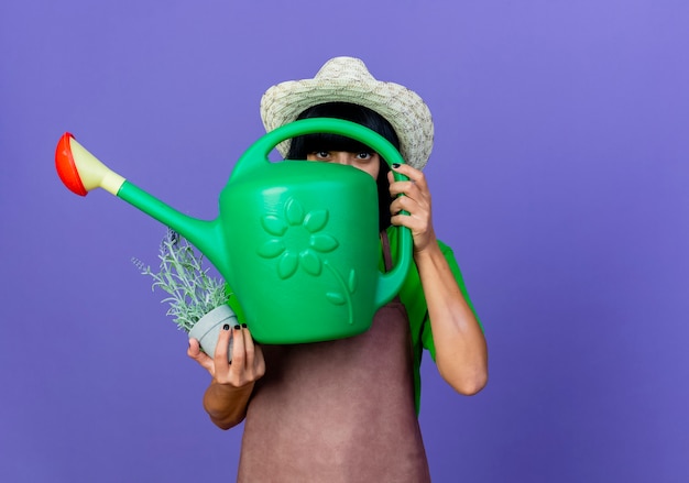 Surprised young female gardener in uniform wearing gardening hat holds watering can and flowerpot