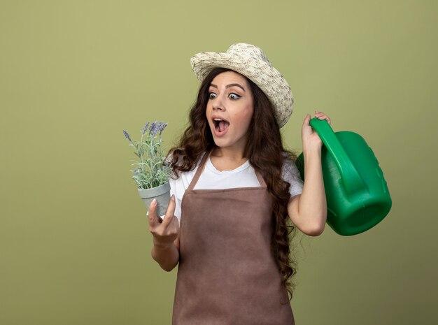 Удивленная молодая женщина-садовник в униформе в садовой шляпе держит лейку и смотрит на цветы в цветочном горшке, изолированном на оливково-зеленой стене