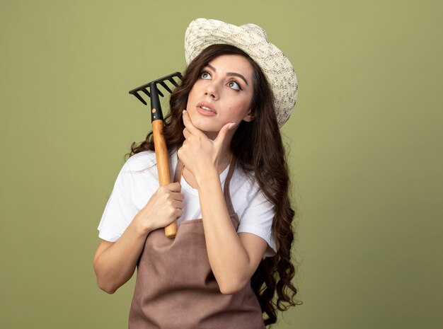 Surprised young female gardener in uniform wearing gardening hat puts hand on chin and holds rake looking up isolated on olive green wall