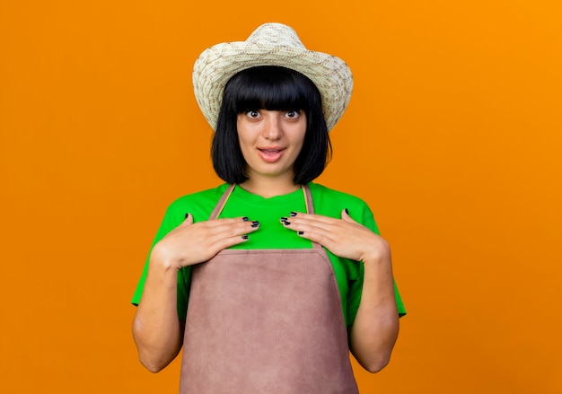 Free photo surprised young female gardener in uniform wearing gardening hat puts hands on chest