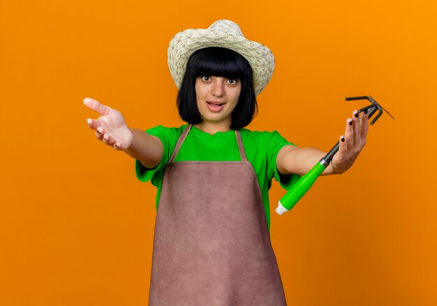 Surprised young female gardener in uniform wearing gardening hat stretching out hands