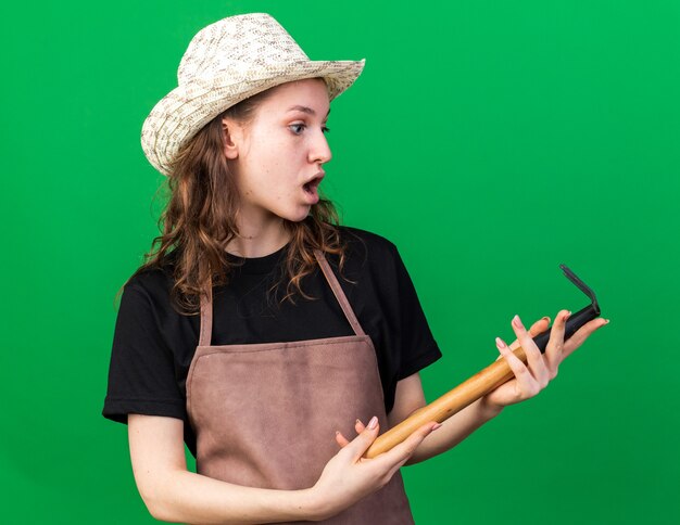 Surprised young female gardener wearing gardening hat holding and looking at rake isolated on green wall