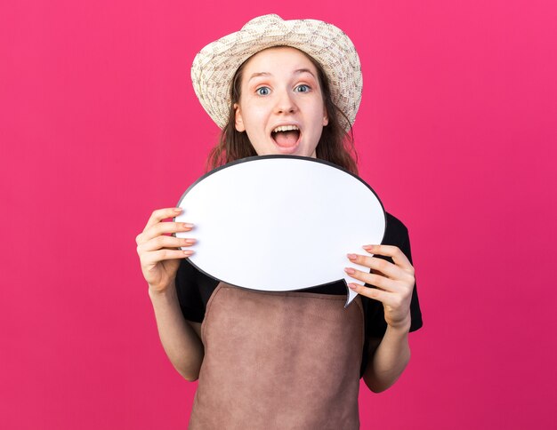 Surprised young female gardener wearing gardening hat holding speech bubble 