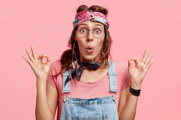 Free photo surprised young female hippie with bugged eyes, makes okay gesture with both hands, dressed in casual outfit, poses over pink wall, demonstrates approval. body language and subculture concept