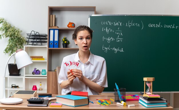 surprised young female math teacher sitting at desk with school supplies holding russian alphabet letter fans looking at front in classroom