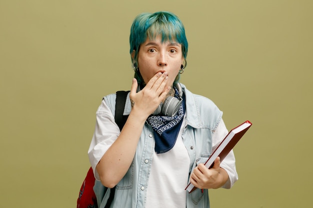 Surprised young female student wearing headphones and bandana on neck and backpack holding note book looking at camera making oops gesture isolated on olive green background