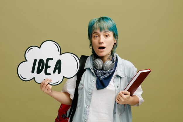 Surprised young female student wearing headphones and bandana on neck and backpack holding note book looking at camera showing idea bubble isolated on olive green background