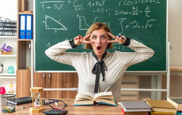 Foto gratuita una giovane insegnante sorpresa si siede al tavolo con gli strumenti della scuola che mostrano un gesto di pace in classe