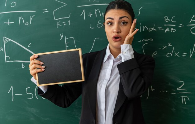 教室でミニ黒板を持って正面の黒板に立っている驚いた若い女教師