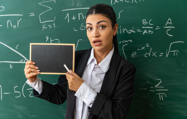 Surprised young female teacher standing in front blackboard holding mini blackboard with stranded for board in classroom