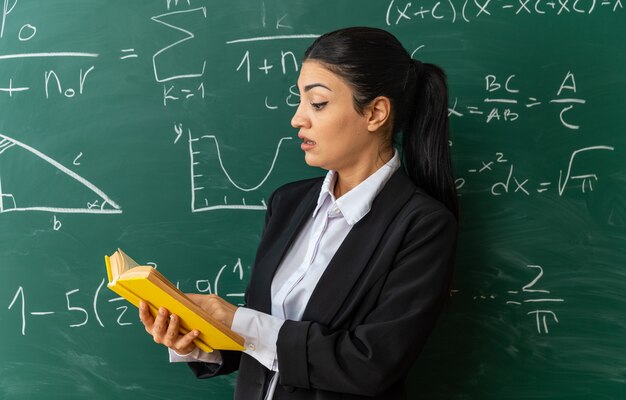 surprised young female teacher standing in front blackboard reading book in classroom