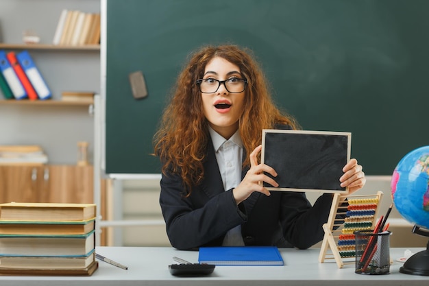 Free photo surprised young female teacher wearing glasses holding mini chalkboard sitting at desk with school tools in classroom