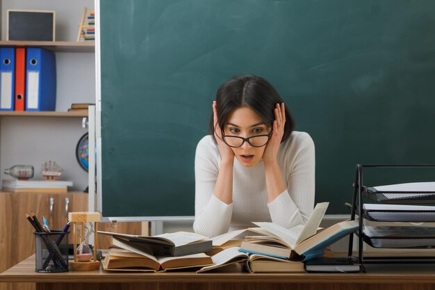 surprised young female teacher wearing glasses reading book sitting at desk with school tools on in classroom