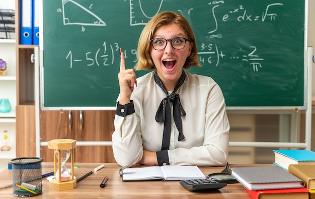 眼鏡をかけている驚いた若い女性教師は、教室で学校のツールポイントを上に向けてテーブルに座っています
