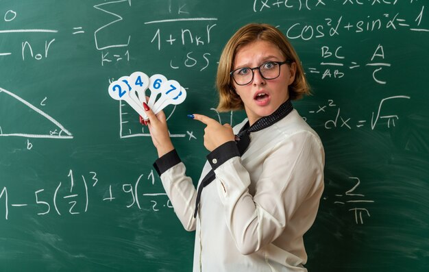 surprised young female teacher wearing glasses standing in front blackboard holding and points at number fans in classroom