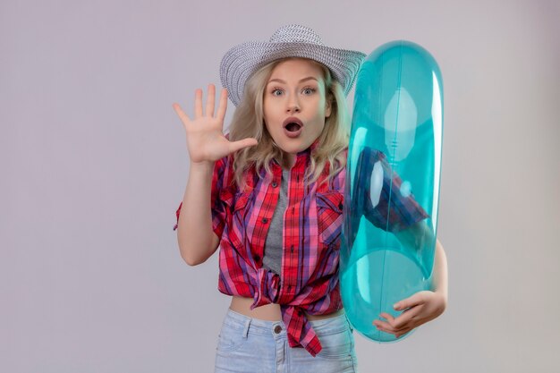 Surprised young female traveler wearing red shirt in hat holding inflatable ring showing stop gesture on isolated white wall