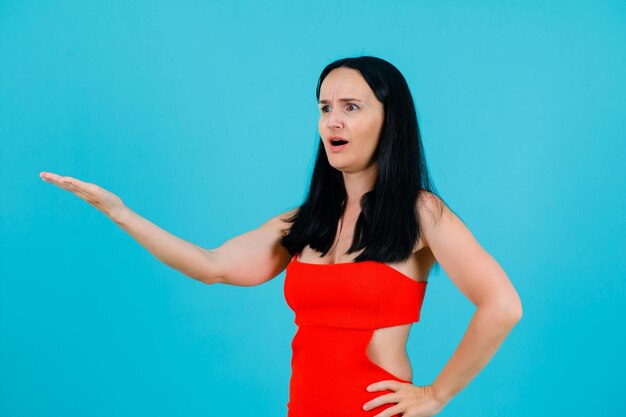 Surprised young girl is extending hand to left and putting other hand on waist on blue background