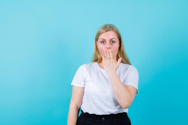 Surprised young girl is holding hand on mouth on blue background
