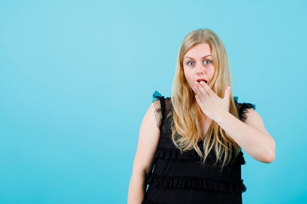 Surprised young girl is looking at camera by holding hand on mouth on blue background