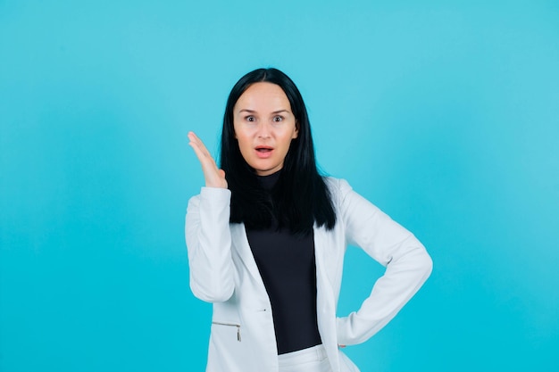 Surprised young girl is looking at camera by holding up hand and putting other on waist on blue background