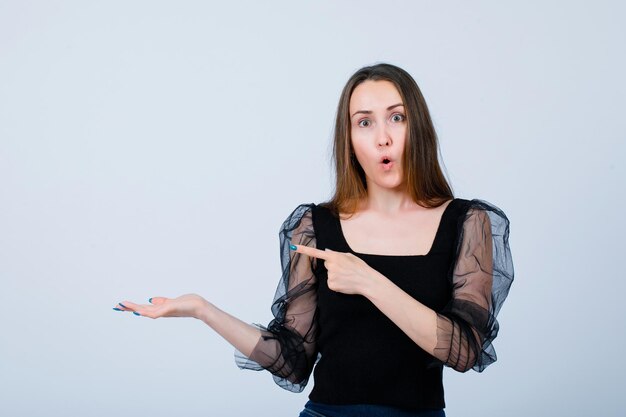 Surprised young girl is looking at camera by pointing left with forefinger on white background