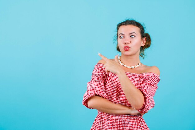 Surprised young girl is looking and pointing left with forefinger on blue background