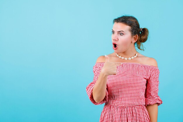 Surprised young girl is looking right and showing herself with forefinger on blue background