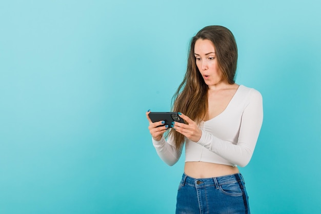 Surprised young girl is looking at smartphone screen on blue background