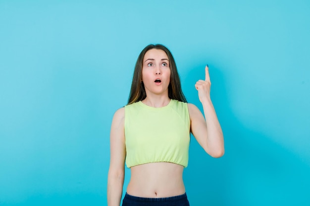 Surprised young girl is looking up and pointing up with forefinger on blue background