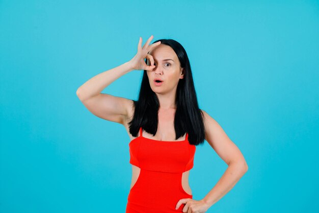 Surprised young girl is making circle fingers around eye and putting other hand on waist on blue background