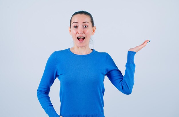 Surprised young girl is opening wide her mouth and pointing right with hand on white background