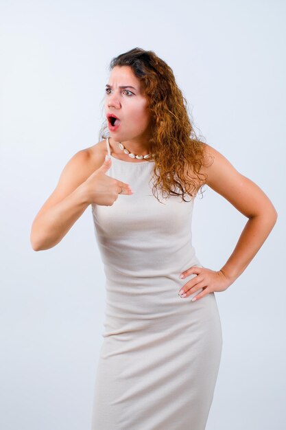 Surprised young girl is pointing herself with forefinger and putting other hand on waist on white background