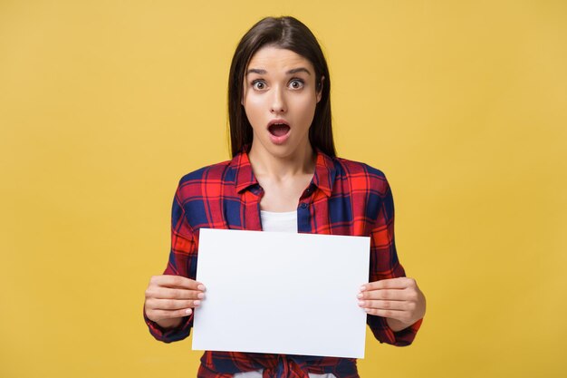 Surprised young girl in red shirt with white placard paper in hands isolated on yellow background
