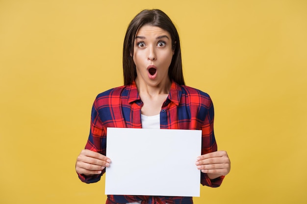 Surprised young girl in red shirt with white placard paper in hands isolated on yellow background