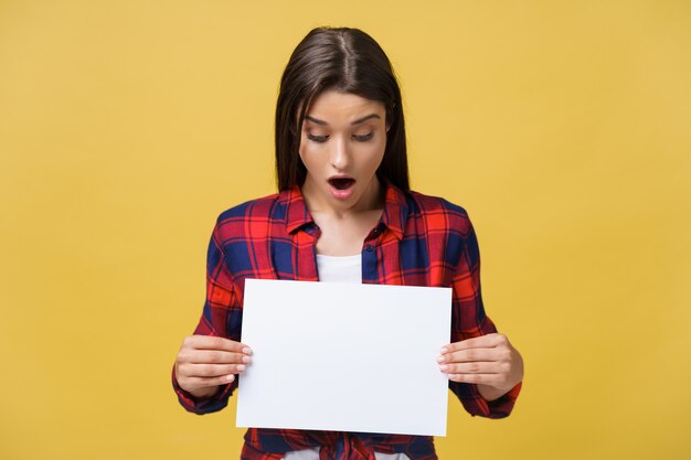 Surprised young girl in red shirt with white placard paper in hands isolated on yellow background.