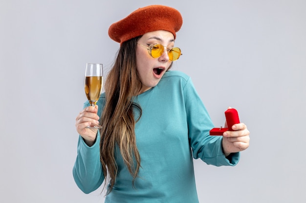 Surprised young girl on valentines day wearing hat with glasses holding glass of champagne with wedding ring isolated on white background