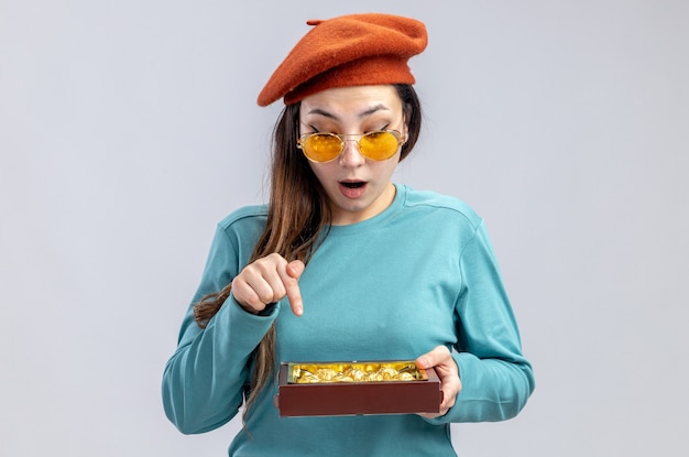 Surprised young girl on valentines day wearing hat with glasses holding and points at box of candies isolated on white background