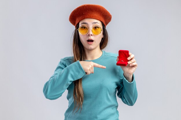 Surprised young girl on valentines day wearing hat with glasses holding and points at wedding ring isolated on white background