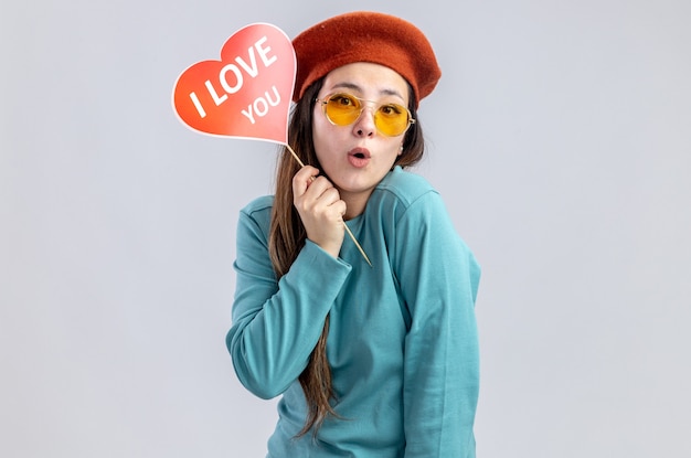 Surprised young girl on valentines day wearing hat with glasses holding red heart on a stick with i love you text isolated on white background