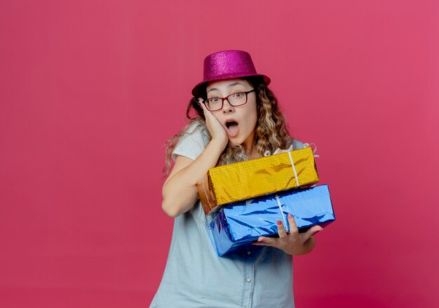 Surprised young girl wearing glasses and pink hat holding gift boxes and putting hand on cheek isolated on pink