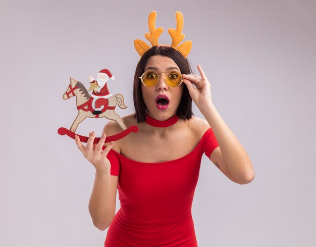 Surprised young girl wearing reindeer antlers headband and glasses holding santa on rocking horse figurine looking at camera grabbing glasses isolated on white background