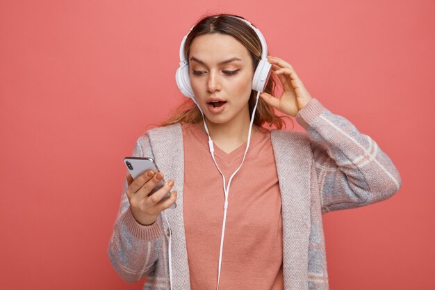 Free photo surprised young girl wearing and touching headphones holding and looking at mobile phone