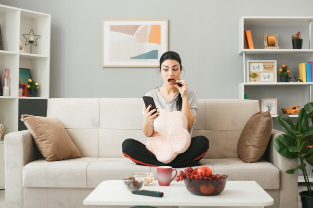 Surprised young girl with pillow holding and looking at phone eat biscuits sitting on sofa behind coffee table in living room