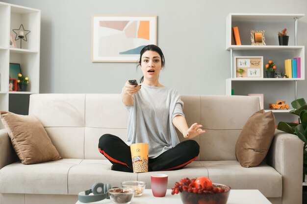 Удивленная молодая девушка с ведром попкорна, держащая пульт от телевизора, сидит на диване за журнальным столиком в гостиной