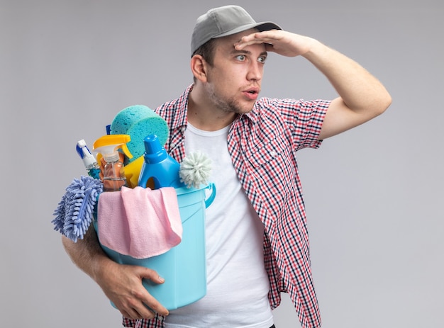 Sorpreso giovane ragazzo pulitore che indossa il cappuccio tenendo il secchio con strumenti di pulizia guardando la distanza con la mano isolata sul muro bianco