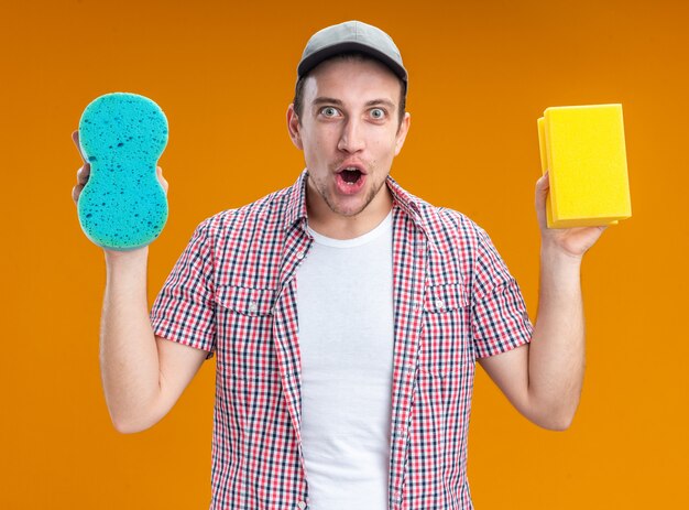 Surprised young guy cleaner wearing cap holding cleaning sponges isolated on orange background