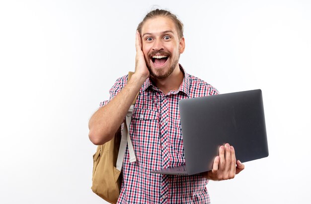 Surprised young guy student wearing backpack holding laptop putting hand on cheek